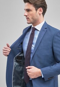 Traje azul con solapa de muesca, bolsillo interior y forro gris. Combinado con una camisa azul claro y una corbata oscura con estampado, sostenido abierto por un hombre.