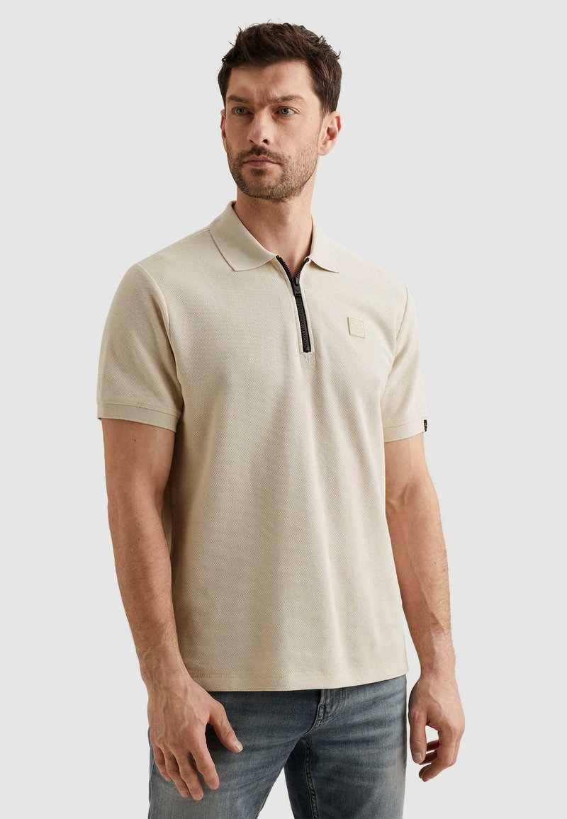 Homme portant un polo beige à manches courtes avec une demi-fermeture éclair noire et un jean bleu clair, debout devant un fond gris clair uni.