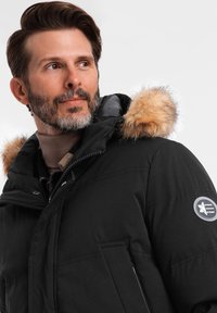 Schwarze Parkajacke mit gefütterter Kapuze aus Pelz, ausgestattet mit einem Reißverschluss und seitlichen Taschen. Strukturiertes Gewebe mit einem Logo-Patch am Ärmel.