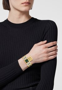 Pulsera en tonos dorado y plateado con esfera cuadrada, dial verde y motivo de león. Presenta un diseño de metal a rayas con colores alternos.
