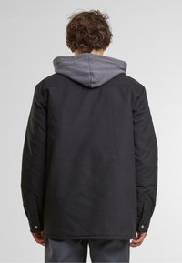 Svart ytterjakke med grå hette; har en avslappet passform, langermet, trykknapper ved mansjettene, og glatt tekstur.