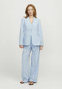Helles blaues Blazer und passende weit geschnittene Hose, beide maßgeschneidert, mit schlanken Revers und subtilen Stickdetails. Getragen mit schlichten Sandalen.