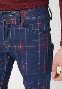Pantalones de mezclilla a cuadros en azul marino con patrones de cuadros rojos y blancos. Presentan cierre de botón, bolsillos laterales y un diseño ajustado.