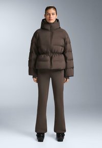 Hnedá bombera s kapucňou, vysokým golierom a prešívaným dizajnom. Spárovaná s rovnakými rozšírenými nohavicami a čiernymi platformovými topánkami.