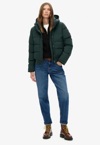 Mørkegrønne pufferjakke med hætte, forreste knapper og quiltet design. Båret over en sort kabelstrikket sweater og blå jeans, parret med brune støvler.