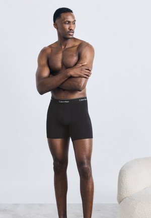 Μυώδης άνδρας με μαύρα ανδρικά μποξεράκια Calvin Klein στέκεται με τα χέρια σταυρωμένα, κοιτώντας προς τα πλάγια, με φόντο ένα απλό φωτεινό τοίχο.