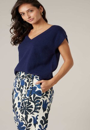 Femme aux longs cheveux ondulés portant un pull à manches courtes bleu marine et un pantalon blanc avec des motifs floraux bleus et noirs, main dans la poche.