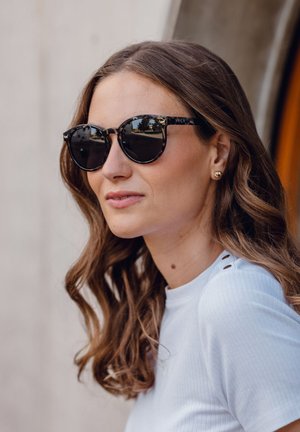 Round, black tortoiseshell sunglasses with dark lenses, featuring a subtle logo on the frame. Worn by a woman with wavy, shoulder-length hair.