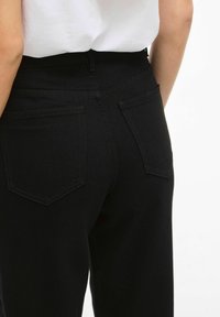 Crne traperice s visokim strukom, s dva stražnja džepa i glatkom teksturom. Jednostavan dizajn s minimalnim detaljima šivanja.