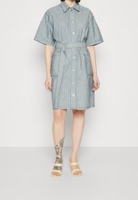 Kvinna iklädd en grå kortärmad knälång klänning med bälte och stora fickor, beige sandaler samt synliga tatueringar på höger arm och vänster ben.