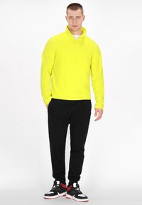 Pullover in pile giallo brillante con colletto alto, abbinato a pantaloni sportivi neri e sneakers alte con accenti rossi e bianchi.