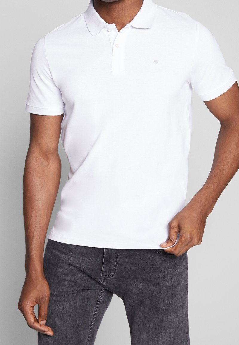 Homme portant un polo blanc uni à manches courtes avec un jean gris foncé, tenant l'ourlet de la chemise d'une main.