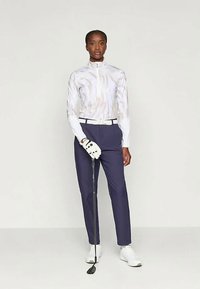 Roupa de golfe com uma parte superior branca com padrão e fecho, calças azul-marinho, cinto branco e luvas brancas, acompanhada de um taco de golfe e sapatos.