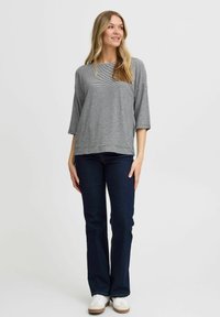 Grau-weißer gestreifter Shirt mit Dreiviertelärmeln, kombiniert mit dunkelblauen Jeans und weißen Schuhen, stehend vor einem schlichten Hintergrund.