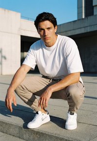 Mladić čučne na gradskom pločniku, nosi bijelu majicu, bež trenirku i bijele tenisice s crnim logotipom pod čistim plavim nebom.