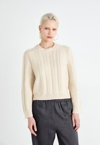 Pull en tricot crème avec un motif en câble texturé, col large et ourlet côtelé, associé à un pantalon gris foncé à coupe relaxée.