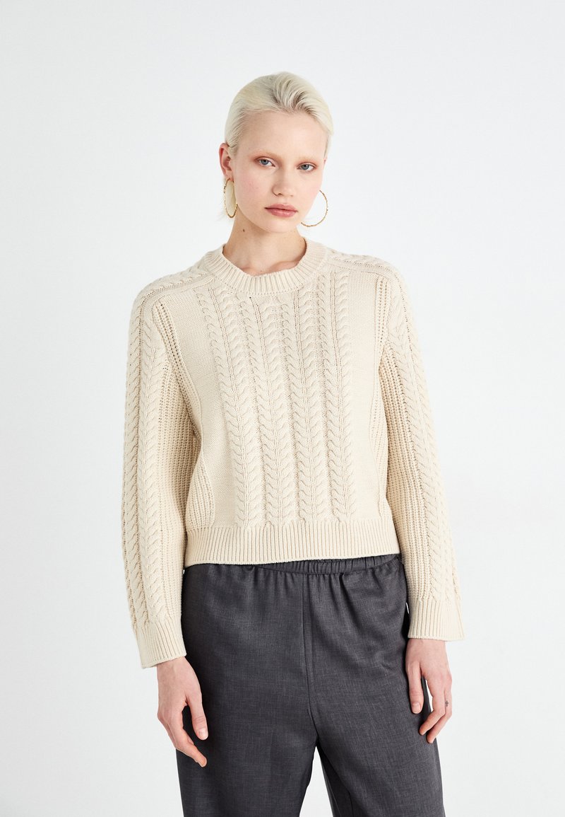 Pull en tricot crème avec un motif en câble texturé, col large et ourlet côtelé, associé à un pantalon gris foncé à coupe relaxée.