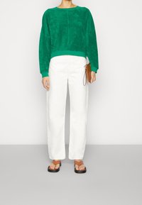 Zelená, strukturovaná mikina s dlouhými rukávy, doplněná volnými bílými kalhotami. Hnědé sandály s černými podrážkami dokončují outfit.