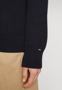 Navy gestrickter Pullover mit einem gerippten Bündchen und einem kleinen Logo-Akzent nahe dem Handgelenk, kombiniert mit beigem Stoff.