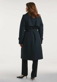 Granatowy płaszcz trenchowy z paskiem w talii i warstwowym tyłem, zestawiony z dopasowanymi czarnymi spodniami i spiczastymi czarnymi szpilkami. Gładka tekstura materiału.