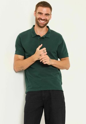 Mann mit kurzen braunen Haaren und Bart, lächelnd, trägt ein dunkelgrünes Polohemd und schwarze Hose, mit Tätowierungen an Händen und Arm.