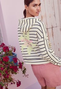 Gestreepte cropped hoodie in geel en zwart, met een groen bloemmotief en tekst op de achterkant. Gecombineerd met roze shorts.