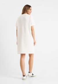 Leichtes knielanges Kleid aus cremefarbenem Stoff mit kurzen Ärmeln und geradem Schnitt, kombiniert mit weißen Sneakers mit schwarzen Akzenten.