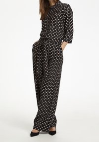 Svart mönstrad jumpsuit med knappstängning fram, trekvartslånga ärmar och bälte i midjan. Har små cirkeldesigner över hela plagg.