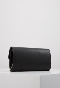 Černá koženka kabelka s cylindrickým tvarem, která má překlopný klopu a detail zlatého řetízku. Hladká textura, minimální zdobení.