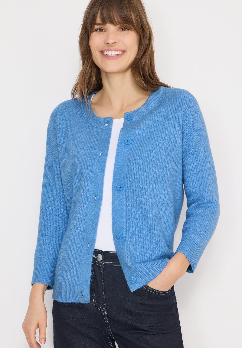 Blue cardigan made of ribbed material, featuring a round neck, long sleeves, and front buttons. Worn over a white top.