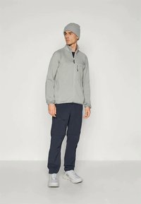 Άνδρας που φοράει ένα ανοιχτό γκρι φούτερ με φερμουάρ, αντίστοιχο σκουφί, σκούρο cargo παντελόνι και λευκά sneakers, στέκεται απέναντι από ένα απλό ανοιχτό φόντο.