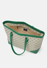 Sac fourre-tout beige avec anses et finitions vertes, orné d’un motif répétant un logo, ouverture en haut laissant apparaître une poche intérieure zippée avec une étiquette de marque.