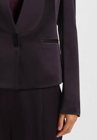 Mørk lilla blazer med en glat tekstur, der har en sjalskrave, en enkelt knaplukning og en sidelomme med en syningdetalje.