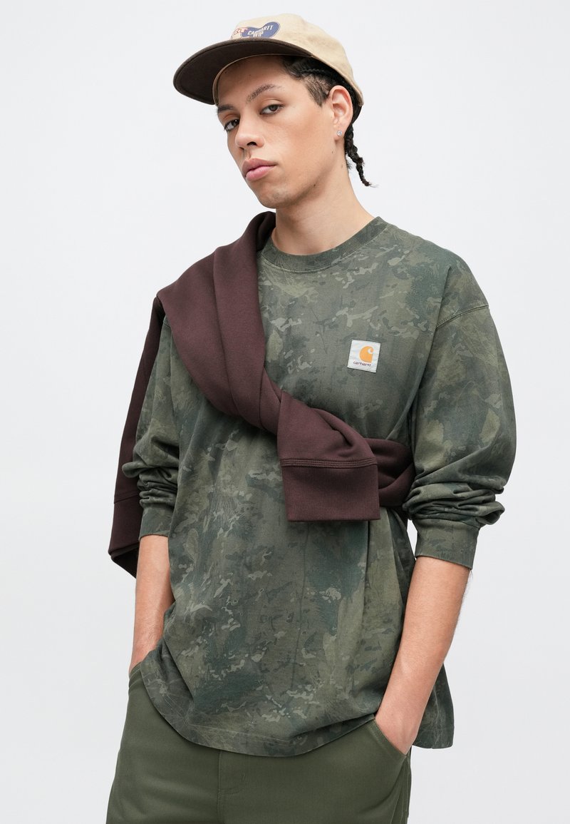 Young person with braided hair wearing a beige cap, camouflage green long-sleeve shirt, and brown sweatshirt draped over shoulders.