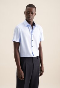 Camicia azzurra chiara a maniche corte con colletto a motivo scuro, apertura con bottoni e texture liscia, abbinata a pantaloni scuri.
