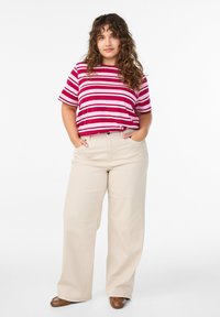 Punaste ja valgete triipudega T-särk, millele on kombineeritud beežid laia säärega püksid. Jalanõud on pruunid. Modellil on lokkis juuksed ja ta seisab käed taskutes.