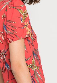 Blouse à fleurs rouges avec des manches courtes bouffantes, ornée d'un imprimé botanique vibrant en jaune, orange et bleu sur un tissu léger.