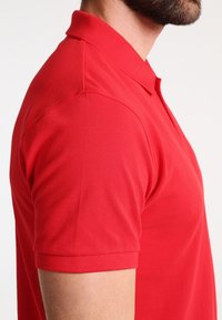 Rød polo skjorte med et tekstureret stof, korte ærmer og klassisk krave. Sidevægten fremhæver form og pasform.