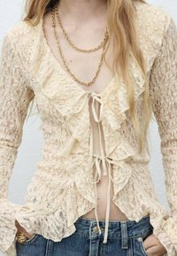 Crèmekleurige kant blouse met gerimpelde voorranden, lange mouwen en een striksluiting. Gecombineerd met hoge taille spijkerbroek shorts en gelaagde gouden kettingen.
