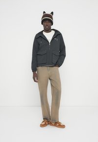 Μαύρο μπουφάν με φερμουάρ και σχέδιο, με δύο μπροστινές τσέπες, μπεζ παντελόνι και καφέ αθλητικά παπούτσια. Πλεκτός σκούφος με διακοσμητικά στα αυτιά σε καφέ και κρεμ ρίγες.