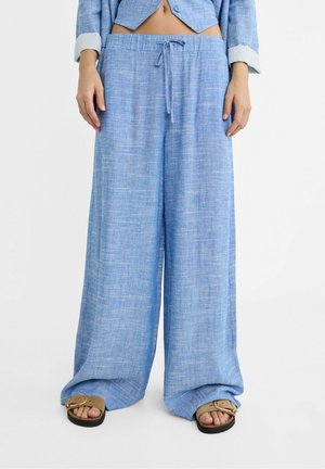 Person trägt hellblaue, weite Hosen mit Zugband, eine verkürzte Jacke und beige Sandalen mit Schnallenriemen vor weißem Hintergrund.