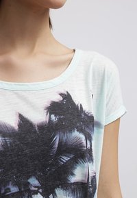 Lyseblå kortærmet T-shirt med rund halsudskæring, der har et mørkt palmetræsgrafik trykt på forsiden mod en blød, tekstureret baggrund.