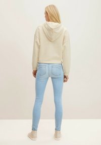 Skuren beige huva med huva, ihopparad med ljusblå slim fit jeans och beige sneakers, visad bakifrån mot en enfärgad bakgrund.