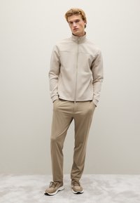 Chaqueta beige con cremallera y cuello alto, combinada con pantalones marrón claro y zapatillas deportivas marrones, todos con texturas suaves y lisas.