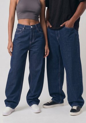 Zwei Personen, die lockere dunkelblaue Jeans und Freizeitschuhe tragen, stehen nebeneinander vor einem einfarbigen Hintergrund.