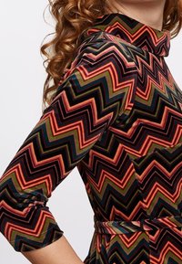 Figursydd klänning med hög krage och trekvartsärmar, tillverkad av mjukt tyg med ett flerfärgat zigzag-mönster i röd, grön och svart.