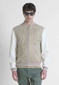 Chaqueta bomber de color beige claro con mangas crema, cierre de cremallera, puños acanalados y logo bordado. Textura de ante suave en el torso.