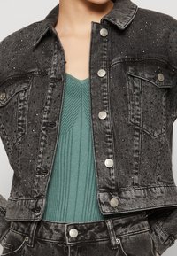 Černá džínová bunda a kalhoty s flitrovými detaily nošené přes modrozelený žebrovaný pletený vršek s výstřihem do V, zobrazené od krku po pas.