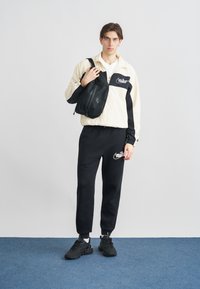 Ανεμοφρακτός της Nike σε κρεμ και μαύρο, συνδυασμένος με μαύρη φόρμα. Το μοντέλο κρατά μια μικρή μαύρη τσάντα με λογότυπο. Μαύρα αθλητικά παπούτσια ολοκληρώνουν την εμφάνιση.