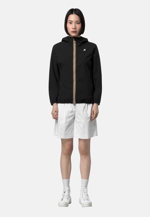 Donna in piedi rivolta in avanti che indossa una giacca nera con cappuccio e logo, pantaloncini bianchi fino al ginocchio, calzini bianchi e stivali bianchi con lacci.